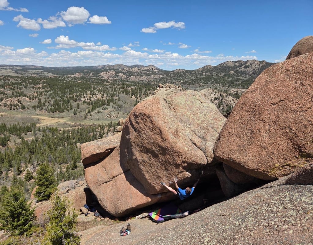 Neil hanging from the underside of a large overhanging pink granite boulder above a wide forested valley