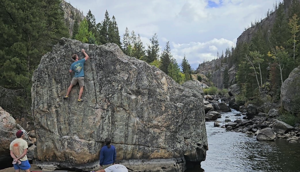 Neil bouldering up the face of a huge granite boulder beside a river in a forested canyon, friends spotting below