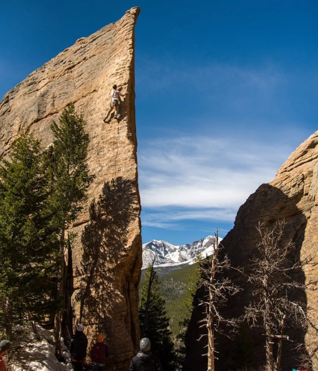 A roped climber high on a tall sandstone spire with snowy peaks and blue sky behind