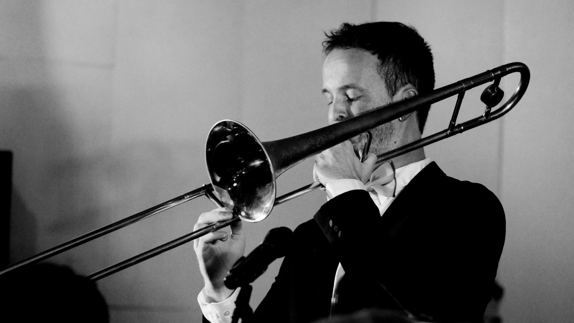 Black and white photo of Neil performing on trombone on stage, wearing a bow tie, eyes closed