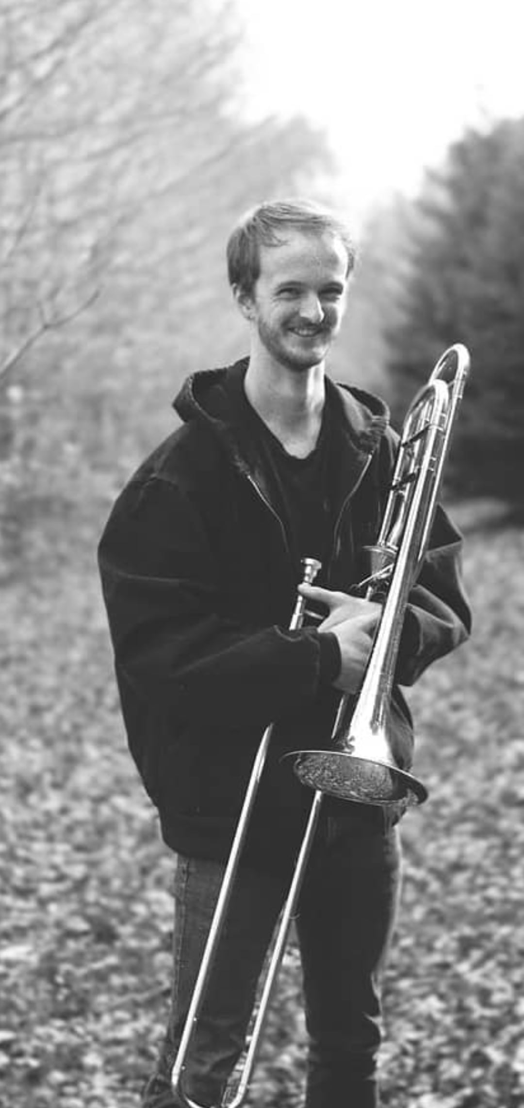Neil smiling outdoors on a leaf-covered path, holding his trombone